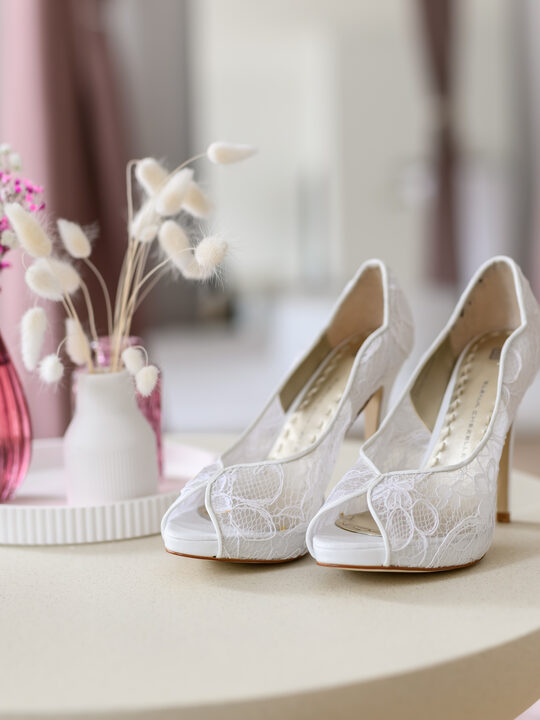 Weiße Brautschuhe mit Spitzenapplikation auf einem Beistelltisch, daneben eine rosa Vase mit Trockenblumen