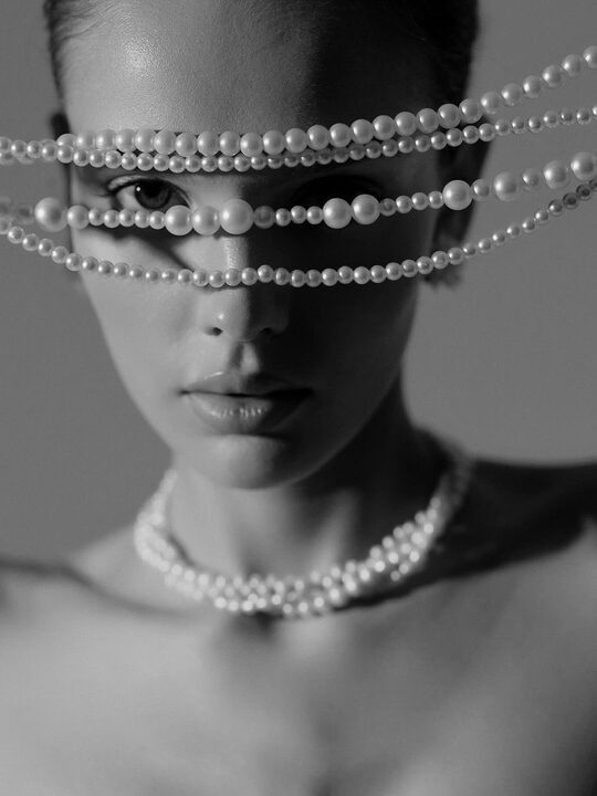 Schwarzweiß-Aufnahme: Perlenketten über den Augen einer Frau, Perlencollier am Hals