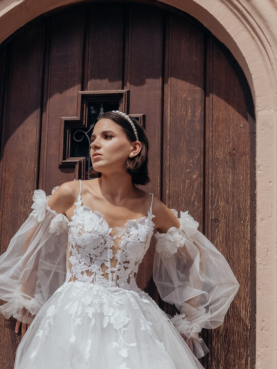 Braut in einem Tüllkleid mit Ballonärmeln vor einem alten Holztor