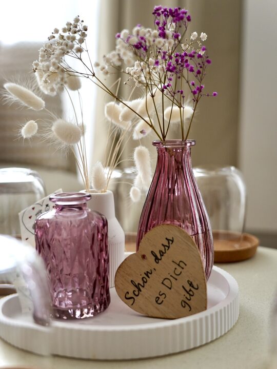Rosa Glasvasen mit Trockenblumen und ein Holzschild mit der Aufschrift „Schön, dass es Dich gibt“
