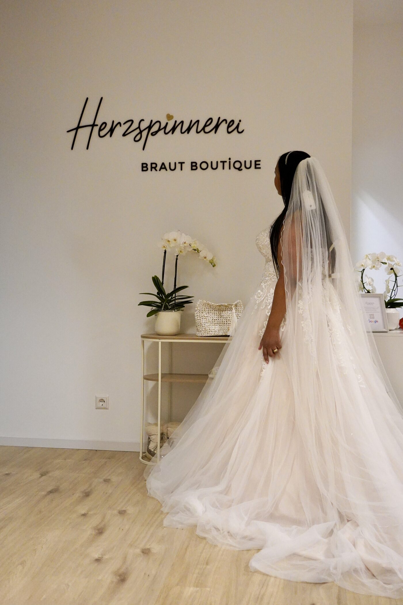 Eine Braut im Tuellkleid mit langem Schleier vor dem Schriftzug der Herzspinnerei Braut Boutique