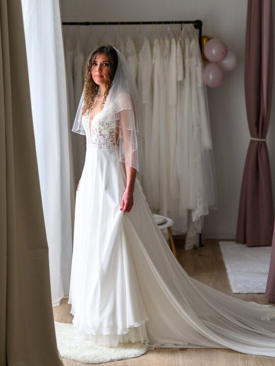 Braut mit Schleier und langer Schleppe vor der Kleiderstange der Boutique